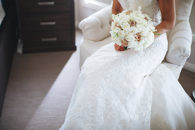 The bride's bouquet of white and champagne roses rests on the lace skirt in a cream armchair.
