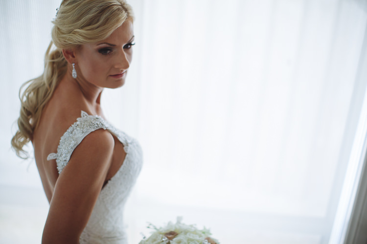 Aggie looks over her shoulder beside sheer curtains, wearing drop earrings and a lace gown.