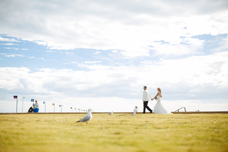 Aggie and Alex walk beside the lawn at Frankston Beach, with gulls beneath a cloudy sky.