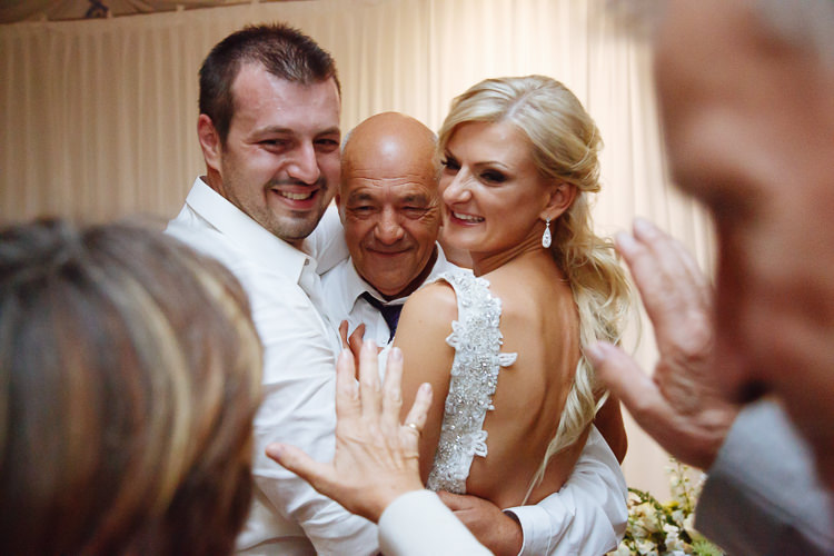 Aggie and Alex embrace a bald older man in a white shirt as nearby guests reach in.