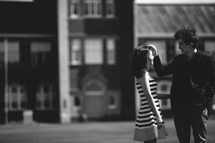 The man sweeps a strand of hair from the woman's cheek, red brick schoolhouse softened behind.