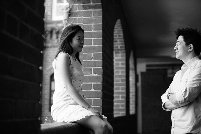 The woman perches on a shaded ledge in white lace while the man watches from the colonnade.