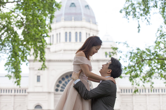 The man lifts the woman toward the dome of the Royal Exhibition Building through spring leaves.