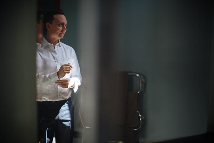The groom threads a cufflink in the slatted window light