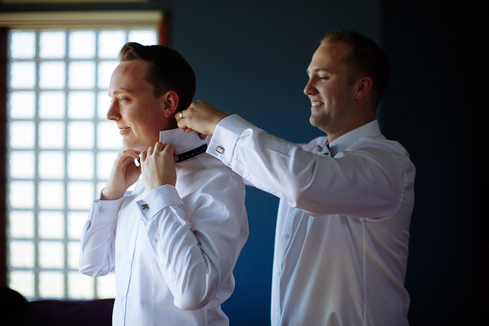 A man in a white shirt fastens the groom's polka dot bow tie from behind at the window.