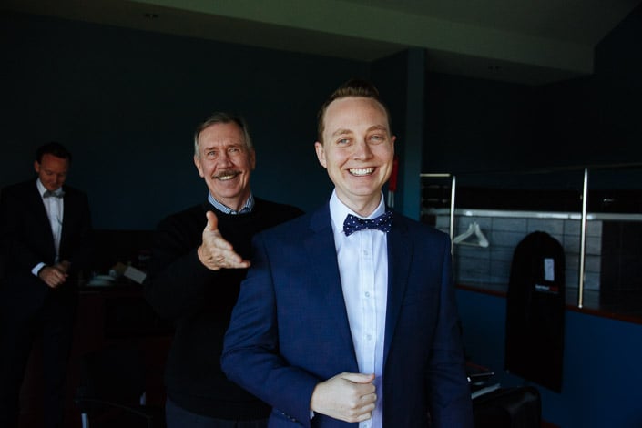 A grey haired man with a moustache reaches an open palm toward camera behind the grinning groom.