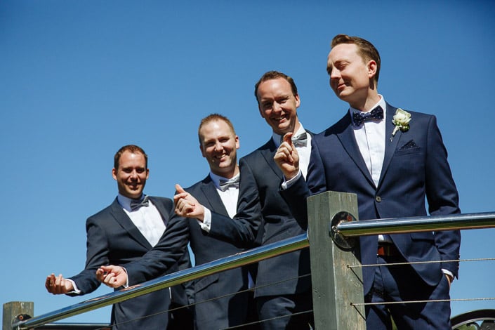 The groom and three groomsmen in navy and bow ties line the rail against a clean blue sky.