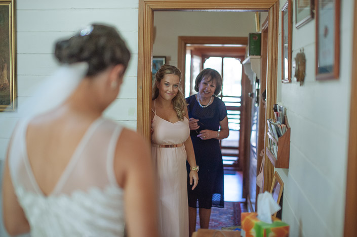 A bridesmaid in blush and the woman in navy and pearls catch the first glimpse from the hallway.