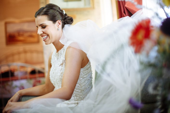 The bride laughs in the armchair as the veil settles behind her, a gerbera vase beside.