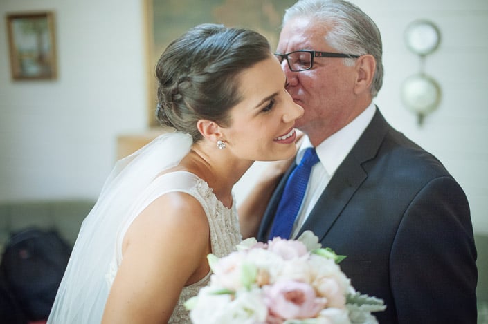 A silver haired man in red framed glasses kisses the bride's temple above the peony bouquet.