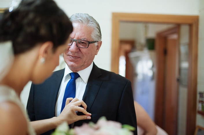 The bride sets the tie straight on the silver haired man in red framed glasses as he blinks.