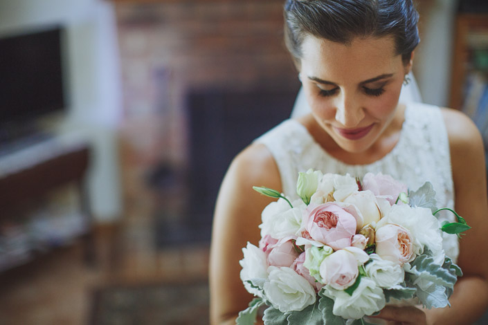 Peonies and dusty miller drawn close beneath the bride's lashes, a brick fireplace behind.