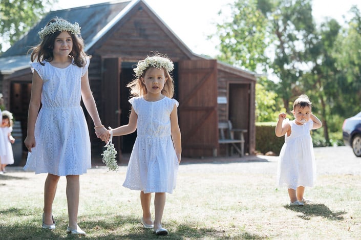 Two flower girls in broderie and baby's breath crowns lead the way, a toddler following behind.