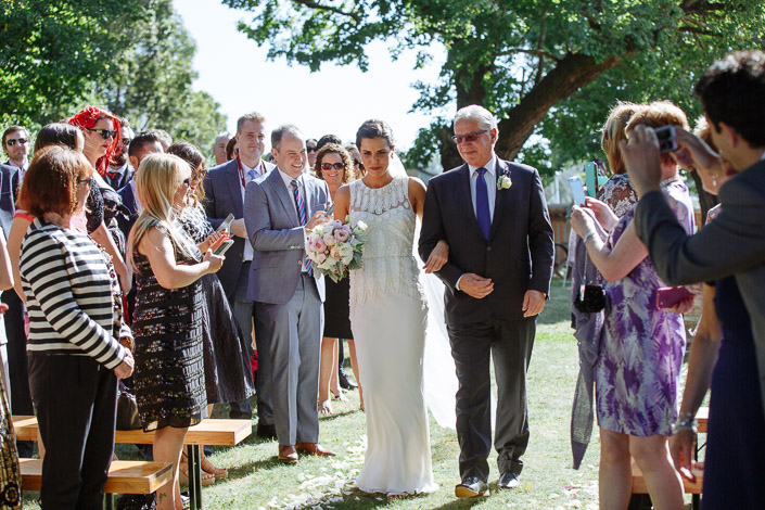 Cameras lift as the bride passes in beaded lace on the arm of the man in red glasses.