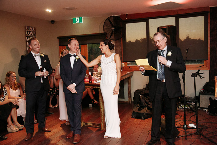 The bride's palm finds the groom's face again as a man in glasses reads from his notes.