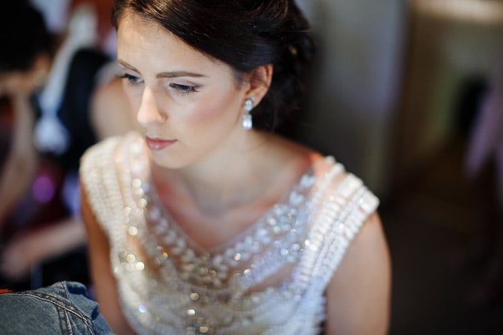 Drop earrings and beadwork glitter as the bride glances down, a denim sleeve at her side.