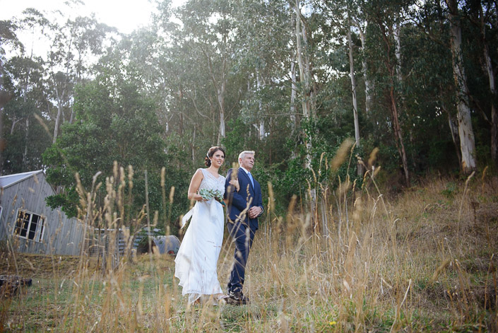 The bride walks down the grassy hill at Pennyroyal Otways Retreat beside a silver-haired man