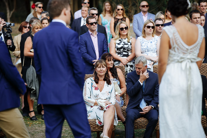 A woman in white florals and a man in navy watch from a wicker chair up front.