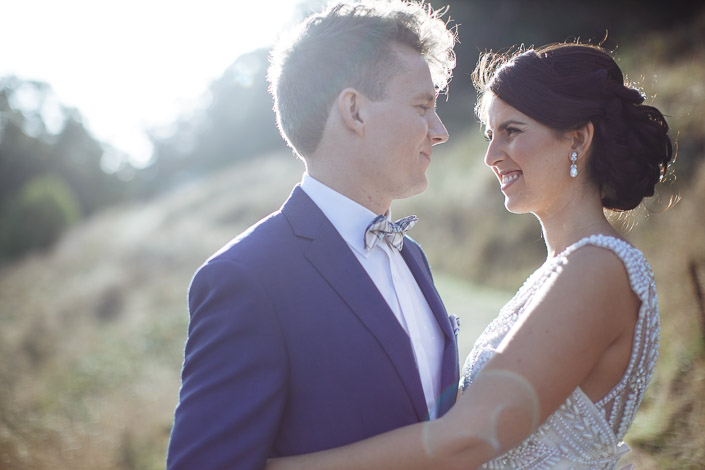 A backlit smile passes between the bride and groom nose to nose in the tall grass.