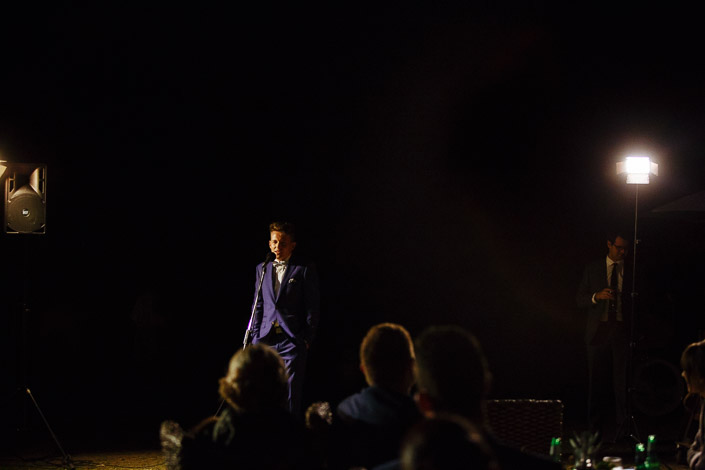 Spotlit at the mic, hands in pockets between the speakers