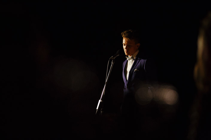 The groom pauses at the mic, spotlit against the dark