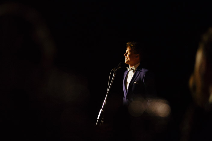 A smile breaks mid-speech under the single beam