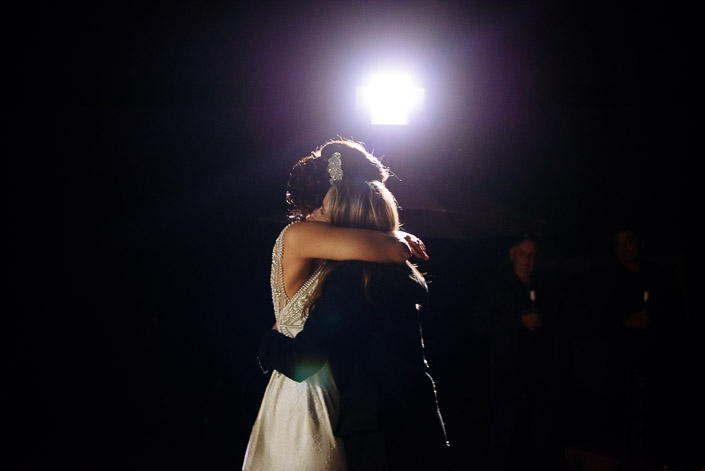 The bride's hug with a blonde guest swallows the spotlight whole, two men watching.