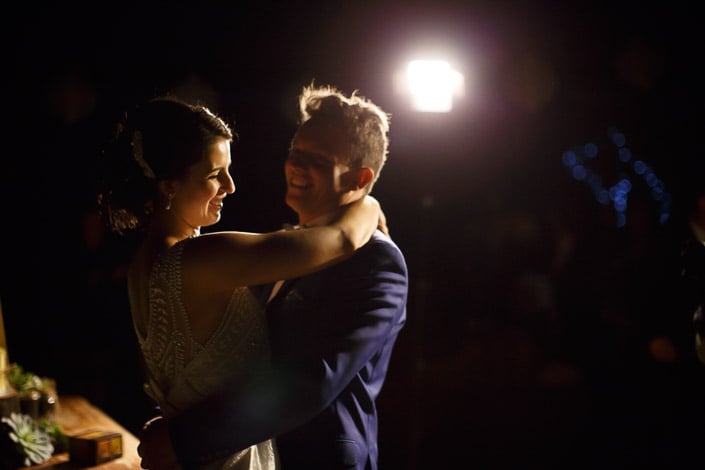 The bride's arms around the groom's neck under the bare bulb glow, fairy lights behind.