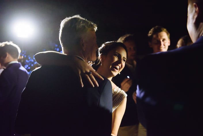 The bride laughs into the shoulder of the silver haired man in a dark suit mid embrace.