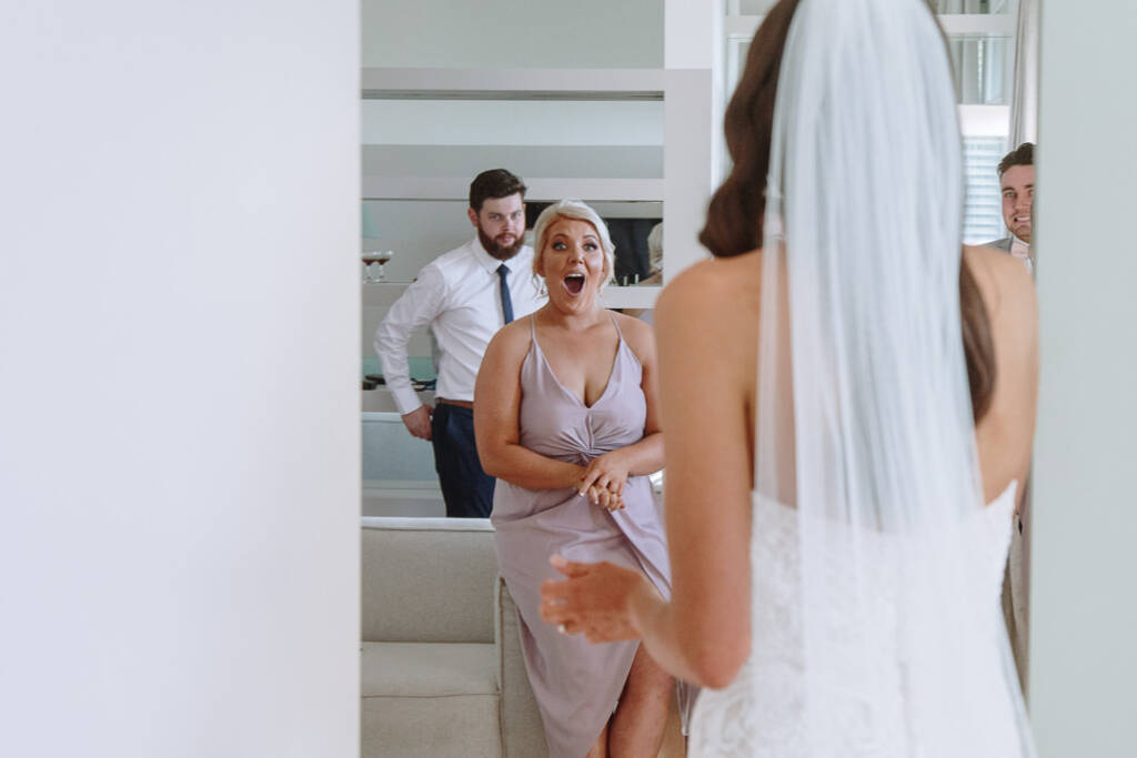 A bridesmaid reacts with an open-mouthed smile on seeing Georgia in her wedding gown.