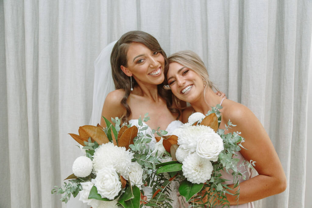 Georgia and a bridesmaid lean their heads together, smiling with their bouquets.