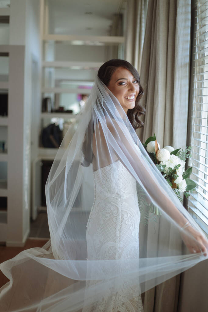 Georgia smiles over her shoulder while holding her veil beside the window at Prince Deck.