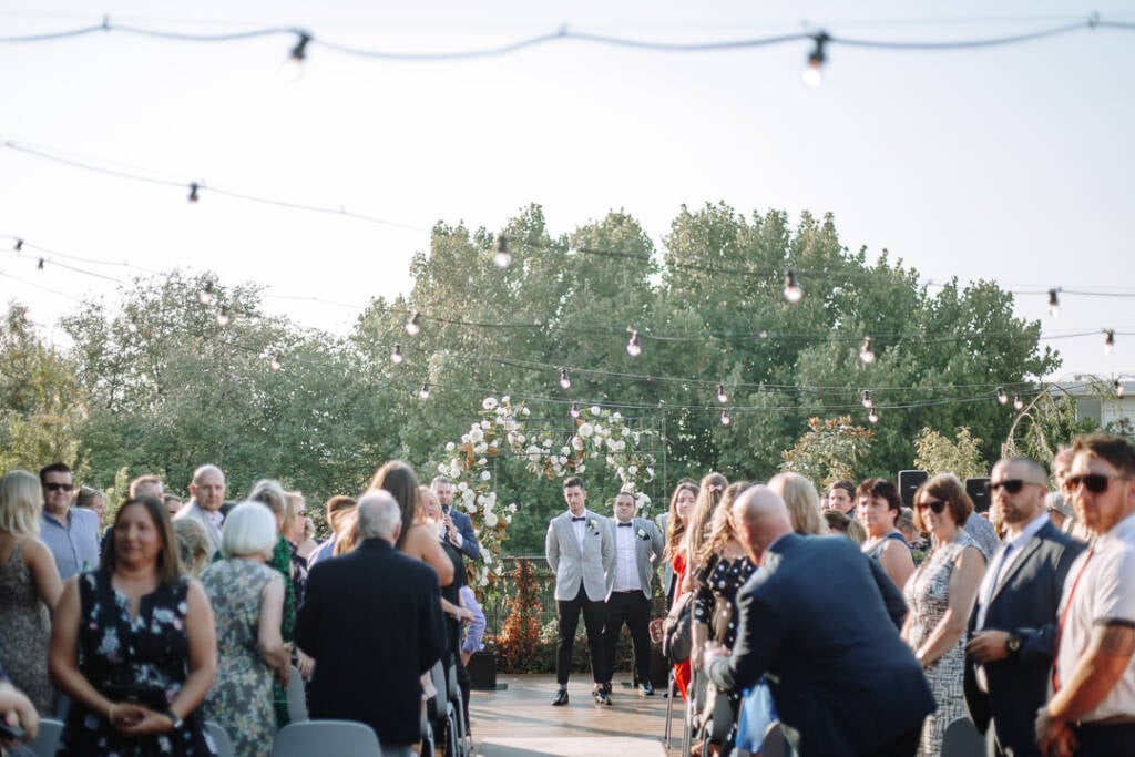 Michael waits beneath the floral arbour at Prince Deck as guests stand beside the timber aisle.