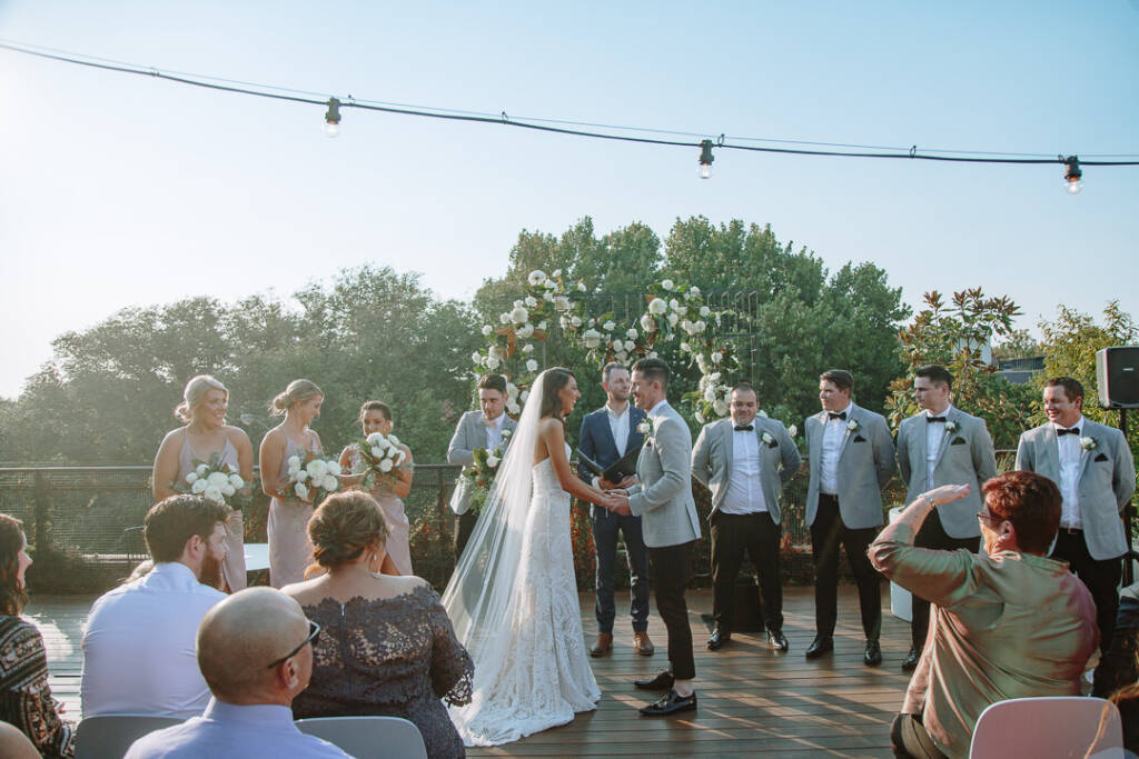 Georgia and Michael hold hands beneath the floral arbour during their St Kilda rooftop wedding at The Prince Deck.