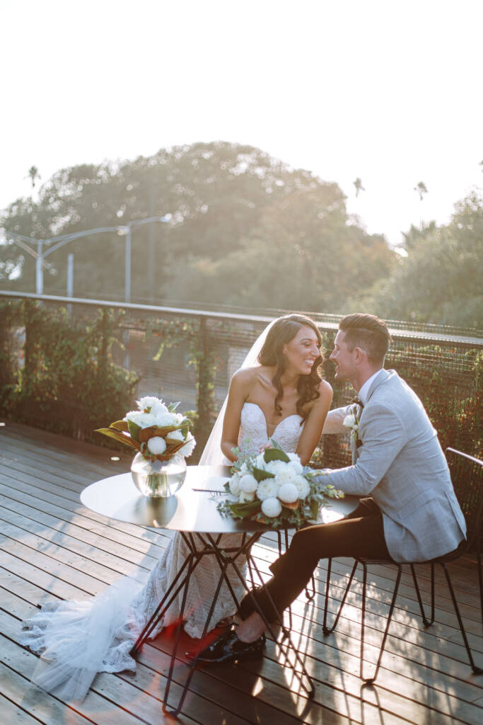 Georgia and Michael lean towards each other at a small table with bouquets and a folder at The Prince Deck.