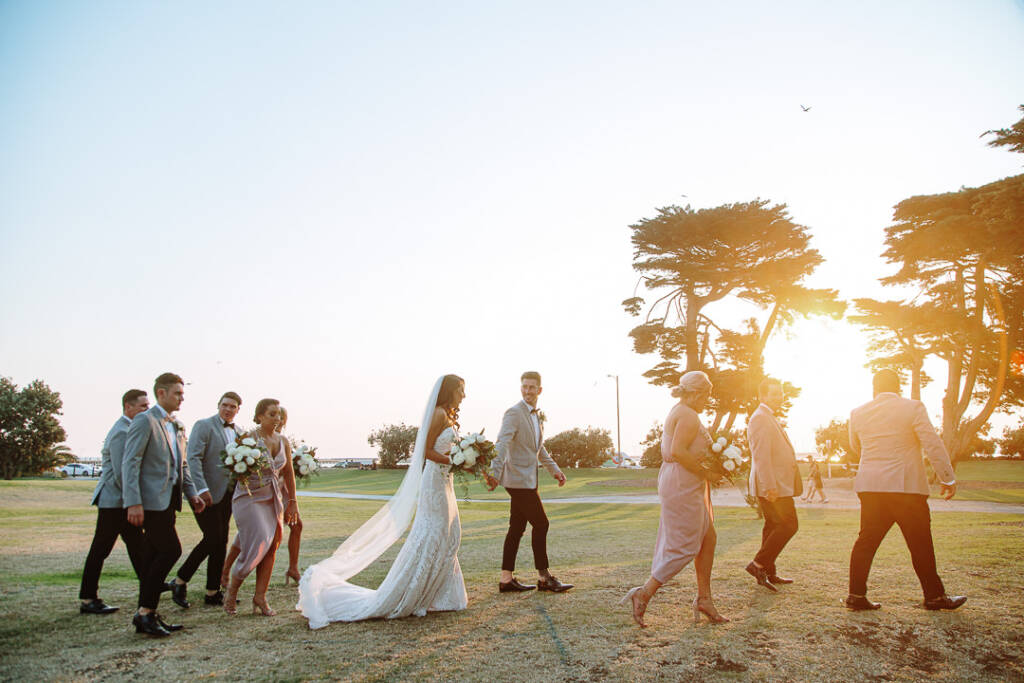 The wedding party walks across the lawn beneath trees at Catani Gardens.