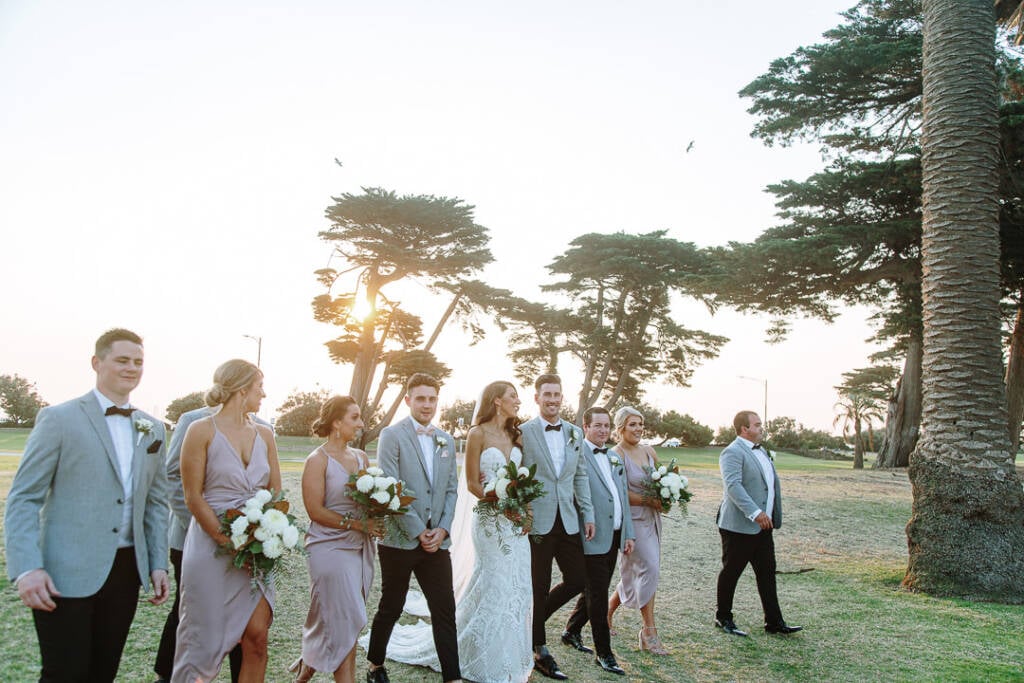 Georgia, Michael and their wedding party walk towards the camera across the lawn at Catani Gardens.