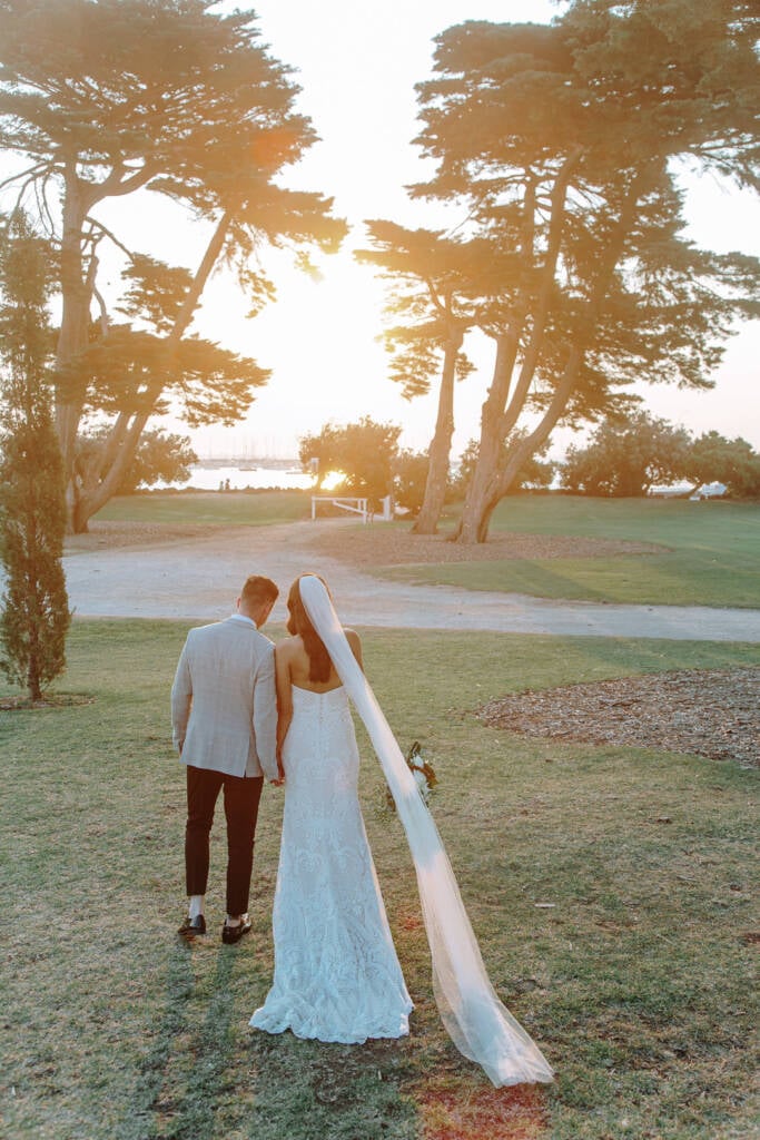 Georgia and Michael hold hands beneath trees at Catani Gardens, with low sunlight beyond the lawn.