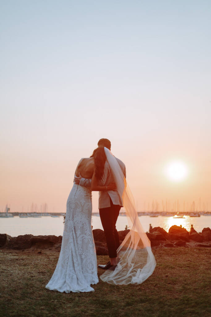 Georgia and Michael embrace during their beach wedding portraits beside St Kilda Beach's rocky edge at sunset.