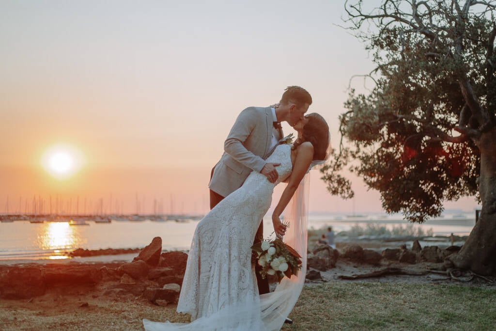 Waterside wedding portrait of Georgia and Michael sharing a dip kiss beside St Kilda Beach at sunset.