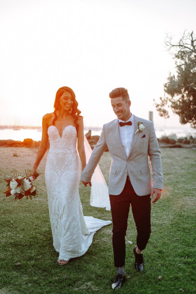 Georgia and Michael walk hand in hand across the grass beside St Kilda Beach at sunset.