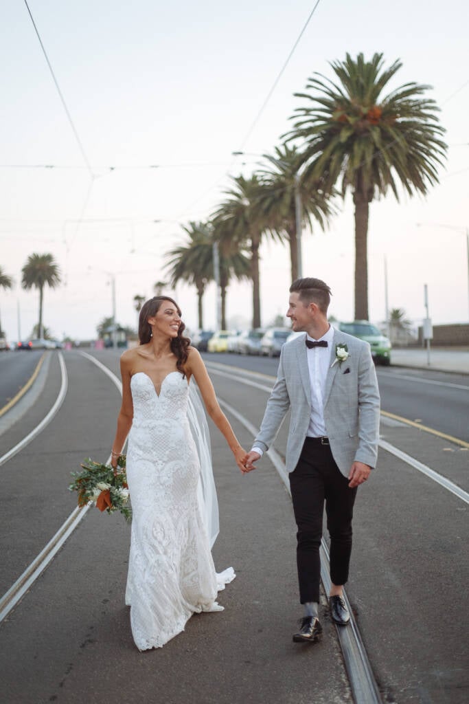 Georgia and Michael hold hands between tram tracks during their urban wedding portraits in St Kilda.