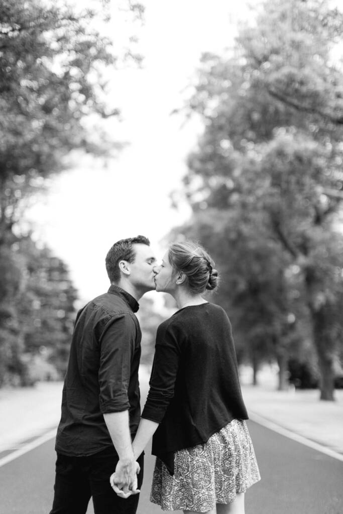 A casually dressed couple kiss on a tree-lined path at Canterbury Gardens.