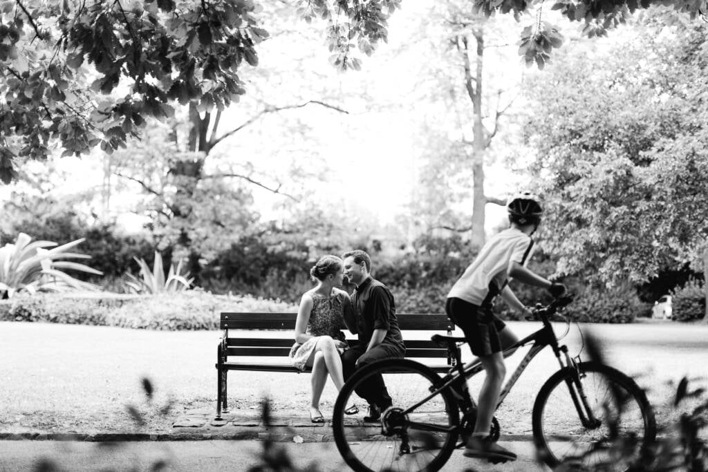 A casually dressed couple sit close on a Canterbury Gardens bench as a cyclist passes in the foreground.