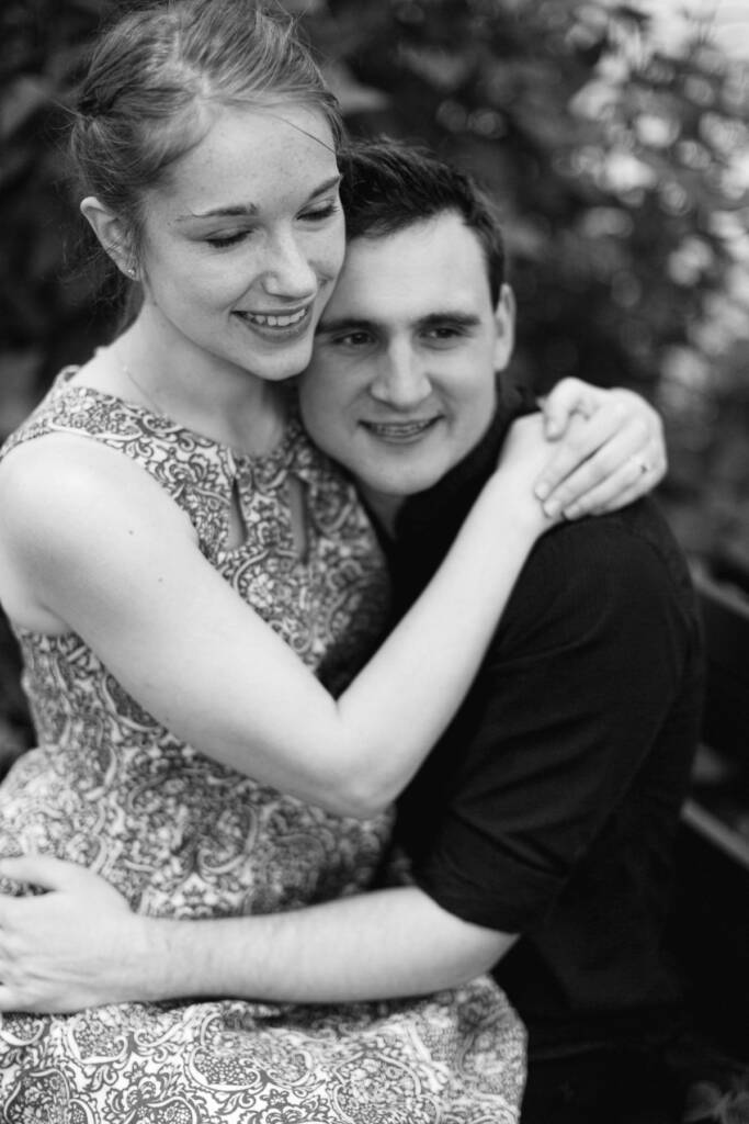 Close black and white of her hugging him from the front, eyes down and smiling, ring showing.