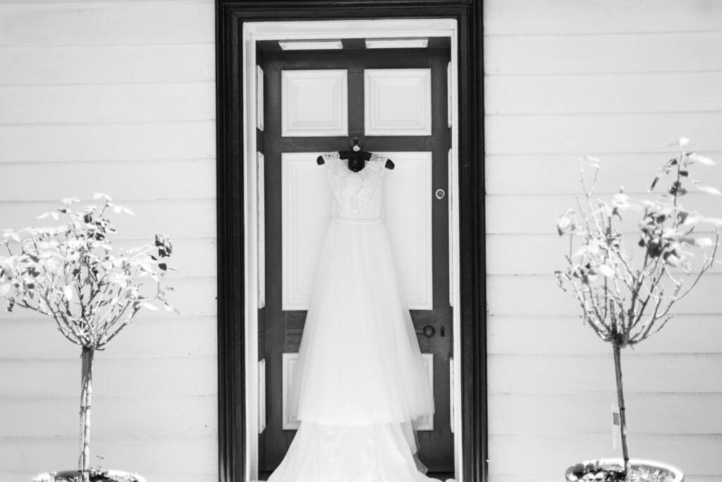 A lace and tulle gown hangs on a dark panelled door in a white weatherboard wall.