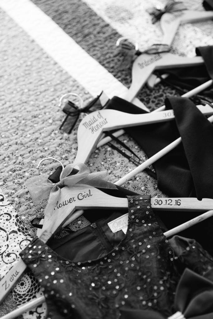 Wooden hangers lettered Flower Girl, Maid of Honour and Bridesmaid lie on a quilt with black dresses.