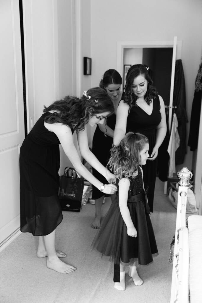 Three bridesmaids in black with flowers in their hair dress the flower girl in a sequinned tulle dress.