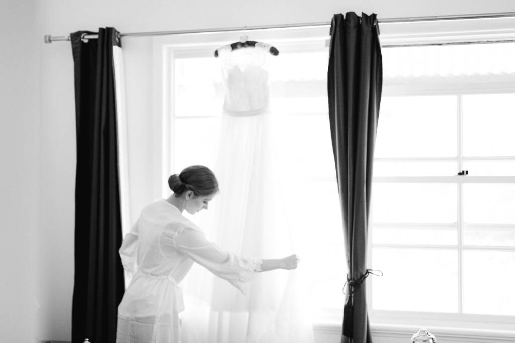 Still in her robe, Sue reaches for the gown hanging backlit in the window between dark curtains.