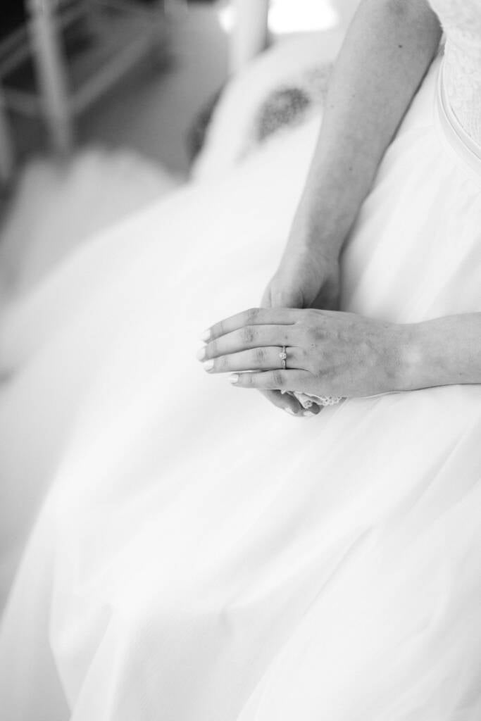 High key close up of Sue's folded hands on tulle, a solitaire ring on her finger.